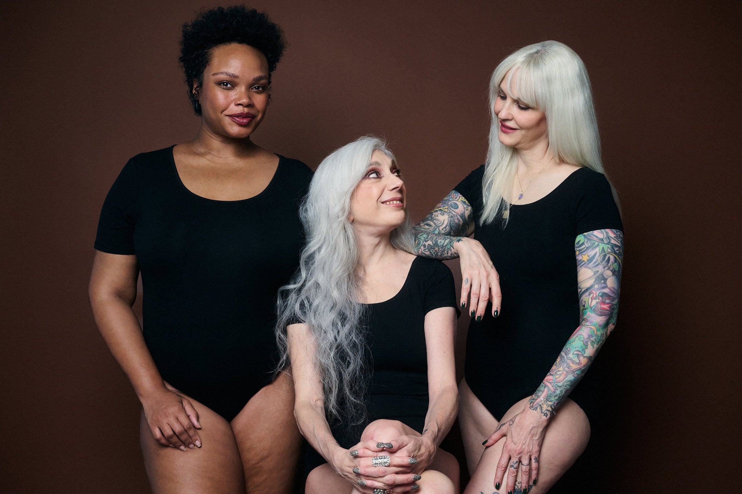 Three women in black bodysuits posing against a brown background